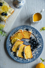 Slices of homemade lemon pound cake decorated with cream cheese frosting, fresh lemon slices and blueberries on vintage blue plate and lemon curd in saucer on linen tablecloth.