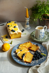 Slices of homemade lemon pound cake decorated with cream cheese frosting, fresh lemon slices and blueberries on vintage blue plate, cake and lemon curd in saucer on linen tablecloth.