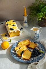 Slices of homemade lemon pound cake decorated with cream cheese frosting, fresh lemon slices and blueberries on vintage blue plate and lemon curd in saucer on linen tablecloth.