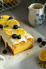 Homemade lemon pound cake decorated with cream cheese frosting, fresh lemon slices and blueberries on baking paper on cutting board and lemon curd in vintage saucer on linen tablecloth.