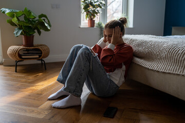 Frustrated sad teen girl child sitting alone on floor being bullied online, having feelings of...