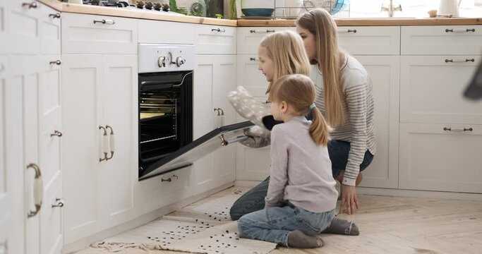 Curious mother and daughters removing baked homemade cookies from microwave and clapping hands in kitchen