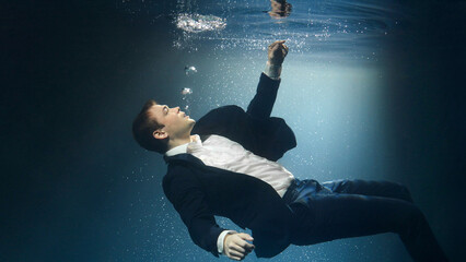 Businessman, a man in a suit, is drowning under water, reaching for the surface of the water.