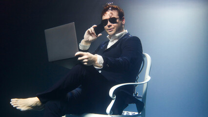 Businessman in a suit is sitting underwater with a laptop and talking on a mobile phone.