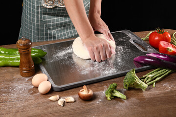 Chef kneading dough on stainless steel cutting board in kitchen with various vegetables next to it,...