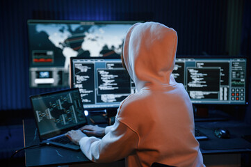 In white hoodie. Young professional female hacker is indoors by computer with lot of information on displays