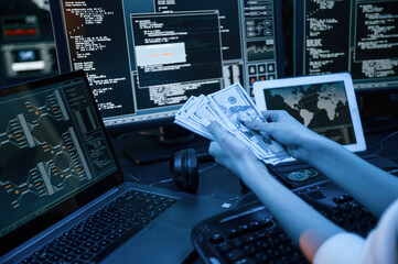 With currency in hands. Young professional female hacker is indoors by computer with lot of...
