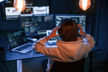 Wearing headphones. Young professional female hacker is indoors by computer with lot of information...