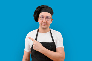 A male chef in an apron on a blue background points his finger at an empty space for menu text.