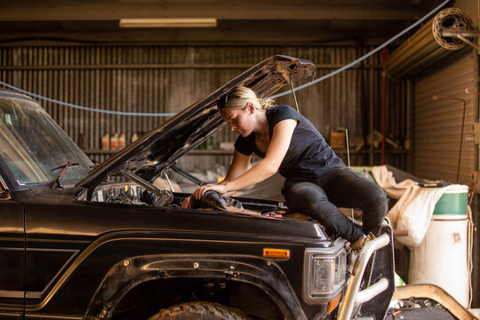 Female mechanic sitting on car hood leaning in repairing the car in workshop