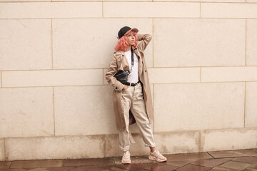 Portrait shooting of a stylish girl with pink hair. Beige shades. Trends of spring and summer 2023. Beige trench cloak with white t-shirt and beige pants. Black small belt bag. Black cap