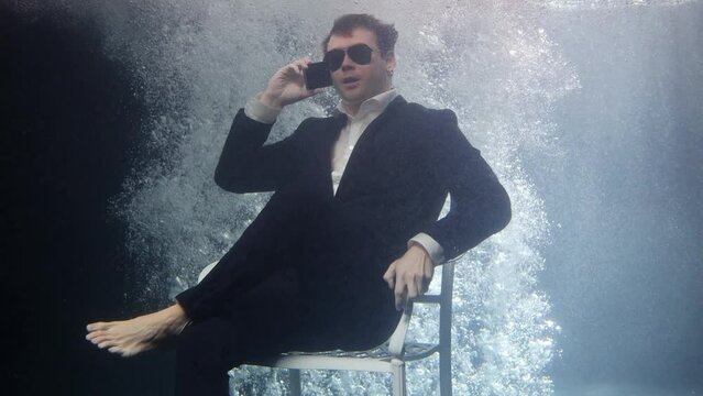 A businessman in a suit and sunglasses is sitting on a chair underwater and talking on the phone.