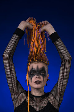 Fine art portrait of beautiful young woman with orange color dreadlocks hairstyle and horror black stage make-up painted on face. Front view, studio shot on blue background. Part of photo series