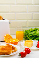 Healthy breakfast in the kitchen. Toaster and bread toast, eggs, tomatoes, freshly squeezed orange juice. Cheese, butter, lettuce. White toaster with healthy food and drinks on table in light kitchen