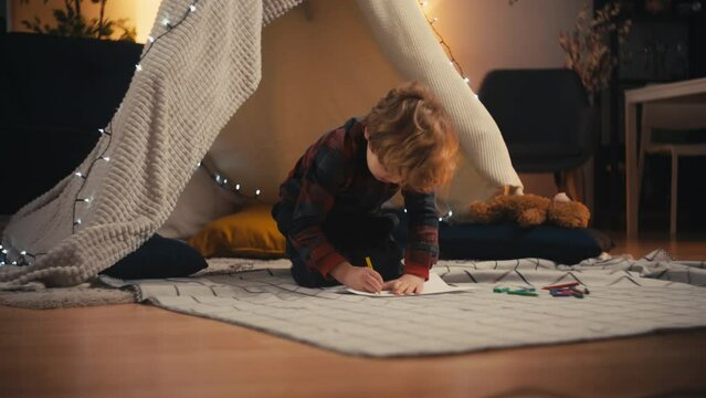 Little boy sitting on the floor in front of blanket fort and drawing, childhood