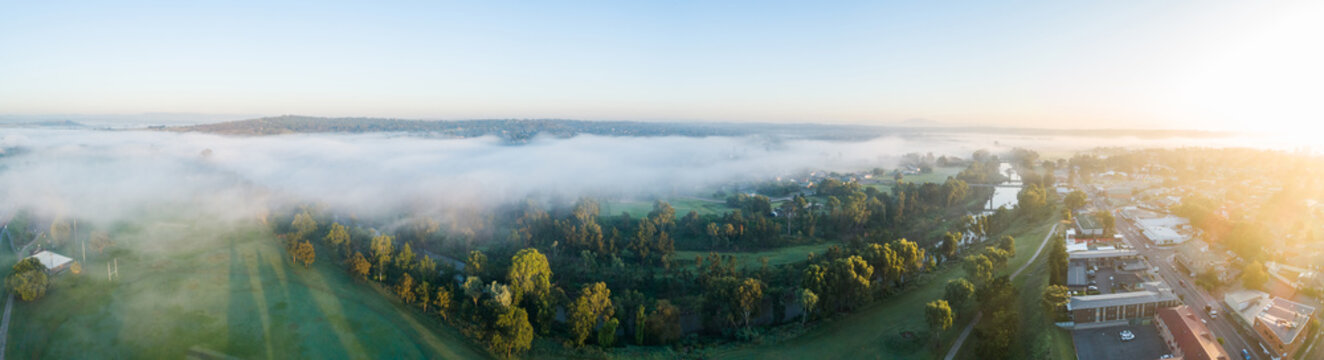 panoramic aerial view of mist over river and farmland beside aussie town at sunrise