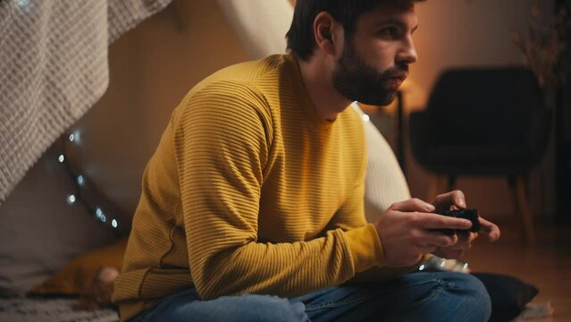 Young man playing video game sitting inside blanket fort, reliving childhood