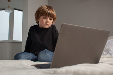 Little boy sitting on bed and looking at laptop. The kid watches cartoons and plays on the computer. 
Toddler learns something new and interesting.