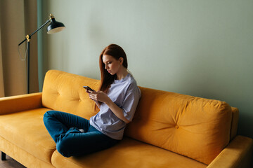Side view of serious redhead young woman looking at smartphone screen sitting on yellow sofa, chatting online, using virtual app on mobile phone, shopping on Internet at home, resting alone.