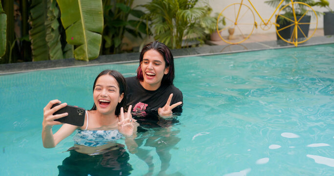 Smiling young women taking selfie in swimming pool female best friend using mobile Indian cute girls making enjoy self portrait photos on smartphone Asian model showing positive emotion v sign hand 