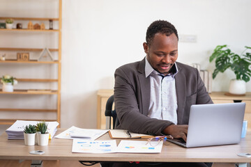 African American businessman analyzing laptop graph paperwork in office holding documents preparing portfolio analysis report Black male analyst doing paperwork in the workplace using the computer.