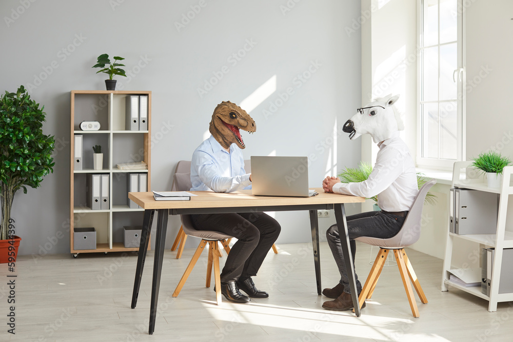 Wall mural Two funny business men wearing animal masks sitting at the desk of their workplace with laptop and having discussion in office, considering new projects or startups, analyzing company.