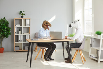 Two funny business men wearing animal masks sitting at the desk of their workplace with laptop and...
