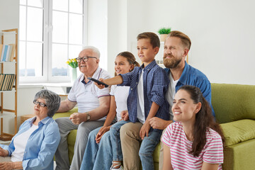 Grandparents, mother, father and two little kids, sister and brother are watching television programs together. Big family is sitting on sofa in front of TV and little boy is using remote control.