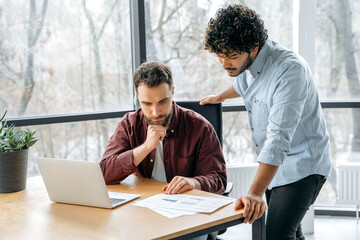 Focused male colleagues of indian and caucasian ethnicity, working together on a project in a modern office, advise each other, share ideas and solutions, planning a product promotion strategy