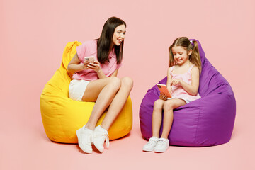 Fototapeta Full body happy woman wear casual clothes with child kid girl 6-7 years old. Mother daughter sit in bag chair hold use mobile cell phone isolated on plain pink background. Family parent day concept. obraz