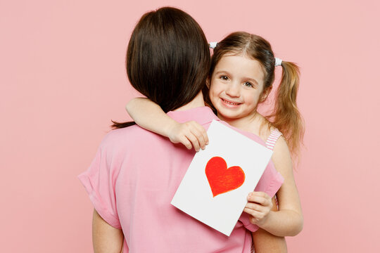 Back rear view woman wear casual clothes with child kid girl 6-7 years old. Daughter give mother postcard with heart, hug and cuddle isolated on plain pastel pink background Family parent day concept