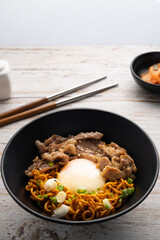 Korean instant noodle with stir fried brisket and onsen egg in black bowl on wooden table.