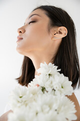 charming asian woman posing with closed eyes near white fresh flowers isolated on grey.