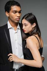 elegant african american guy in black blazer embracing sensual asian woman looking away isolated on grey.