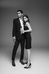 full length of interracial couple in elegant and stylish attire standing with champagne glasses on grey background.
