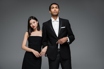 young interracial couple in elegant formal wear looking at camera isolated on grey.