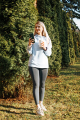 Fototapeta premium Smiling slim young blonde woman using smartphone in park, lifestyle. Cheerful caucasian female student holding phone and looking away on sunny day outdoors