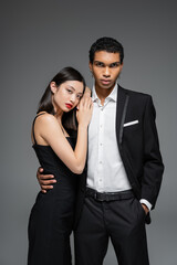 elegant african american man standing with hand in pocket and hugging sensual asian woman with red lips isolated on grey.