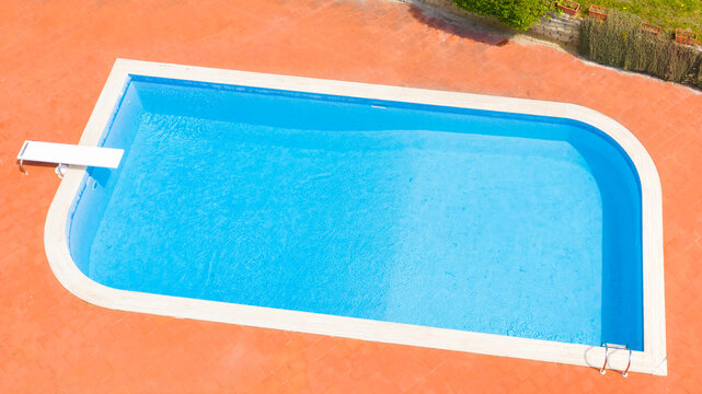 Aerial view of a rectangular swimming pool with diving board, belonging to a large villa. Around the water there is a stone floor. The pool is empty and there is nobody in the garden.