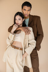 trendy african american man embracing asian woman in pantsuit and looking at camera isolated on beige.