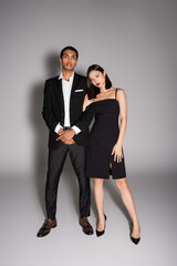 full length of interracial couple in elegant formal wear looking at camera on grey background.