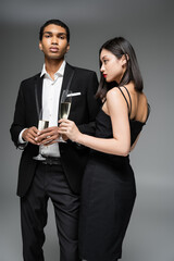 young interracial couple in black elegant clothes posing with champagne glasses isolated on grey.