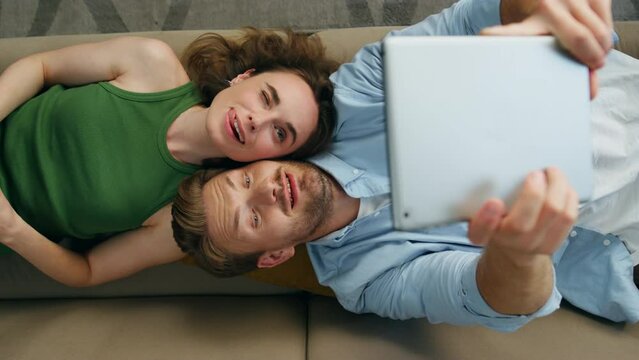 Funny couple taking selfie laying couch top view. Happy man photographing tablet