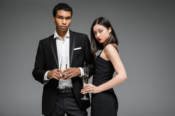african american man in black suit and asian woman in strap dress holding champagne and looking at camera isolated on grey.
