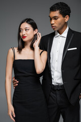 african american man in black blazer hugging waist of elegant asian woman smiling and looking away isolated on grey.
