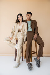 full length of multiethnic couple in stylish pantsuits posing near stools on beige background.