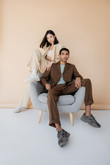 full length of interracial couple in stylish pantsuits posing near armchair on beige background.