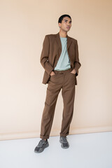 full length of african american man in trendy suits standing with hands in pockets and looking away on beige background.