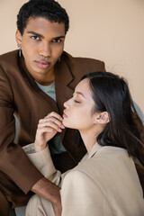 stylish african american guy with piercing looking at camera near young asian woman with closed eyes isolated on beige.