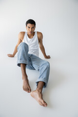 full length of stylish african american man in blue jeans and tank top sitting and looking at camera on grey background.